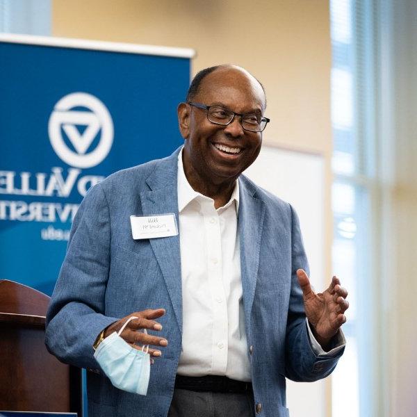 William "Bill" Pickard smiles and gestures with his hands in front of a GVSU vertical banner